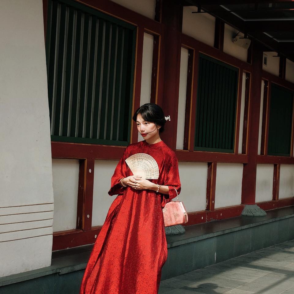 A woman wearing a red traditional dress holds a fan while standing in front of a building with red and green details.