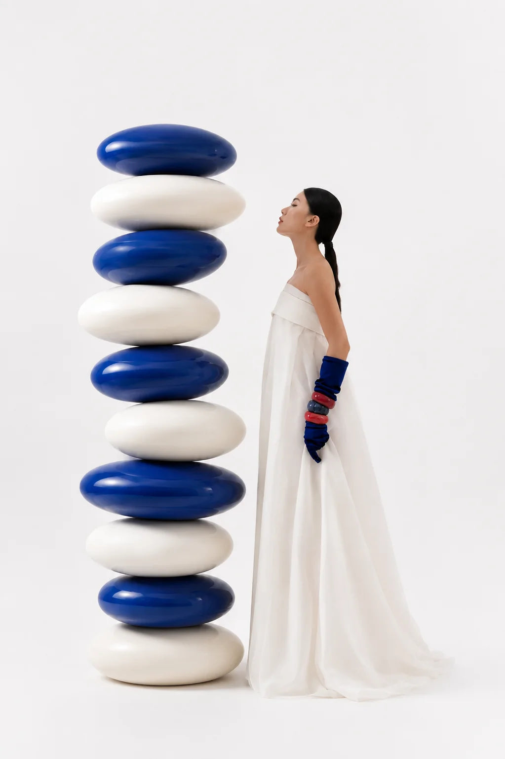A model in a white strapless gown and cobalt gloves stands with eyes closed beside a tall column of stacked cobalt and white stones